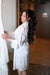 bride in a white silk robe with lace sleeves admiring her wedding gown, styled with voluminous dark wavy hair and romantic bridal makeup by Karysha Beauty