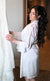 Bride in a white lace robe admiring her wedding dress, styled with elegant bridal makeup and long dark wavy hair by Karysha Beauty